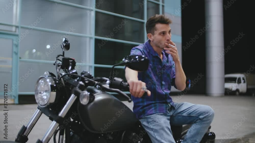 Portrait of man motorcyclist smokes a cigarette while sitting on a motorcycle at night street Portrait of man motorcyclist smokes a cigarette while sitting on a motorcycle at night street