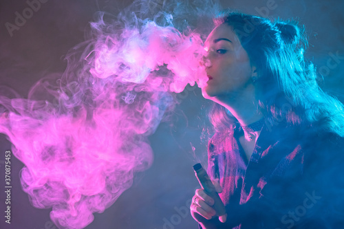 Vaping. Girl with an electronic cigarette. Portrait of a vaper in the smoke. Student exhales a lot of smoke. Woman is engaged in vaping. Young woman holds a vape device. Concept - gift for the vaper