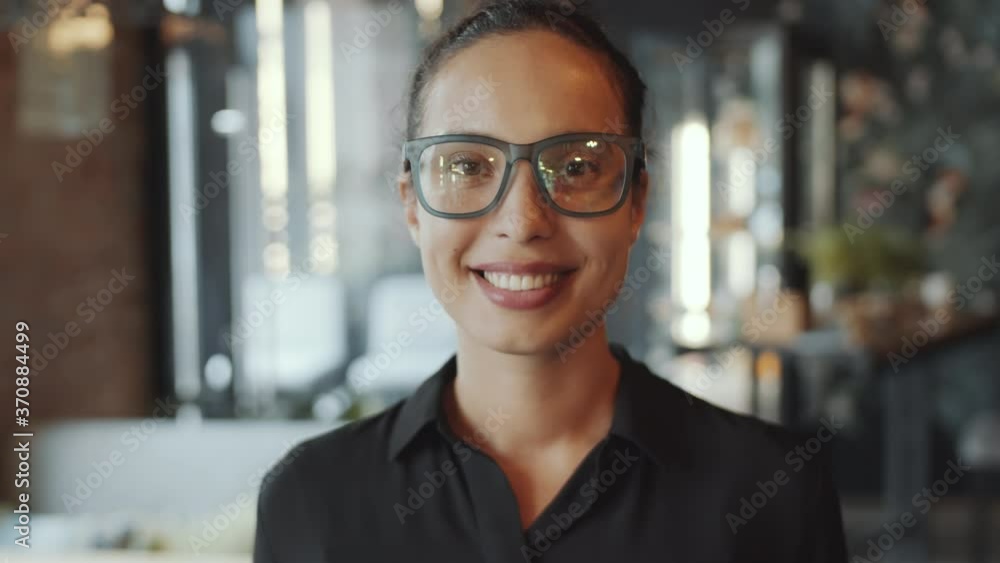 Chest up shot of beautiful mixed-raced businesswoman in elegant black shirt and glasses looking at camera and smiling while standing in cafe Chest up shot of beautiful mixed-raced businesswoman in elegant black shirt and glasses looking at camera and smiling while standing in cafe