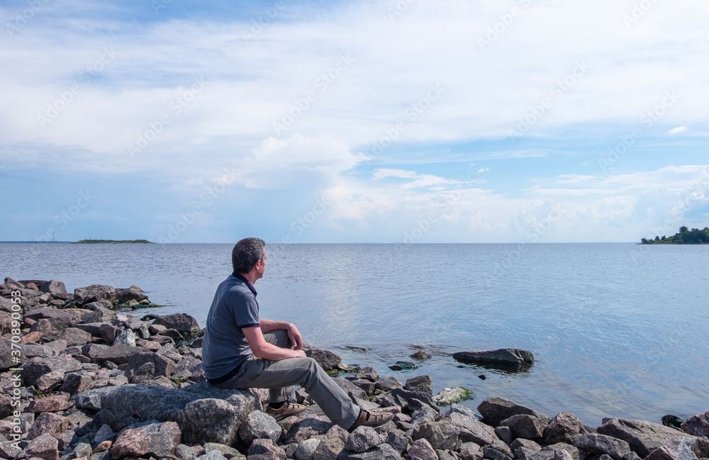 A lonely middle-aged man sits on a rocky beach on a summer day and looks thoughtfully into the distance. Loneliness, unity with nature concept. Horizontal orientation, selective focus.