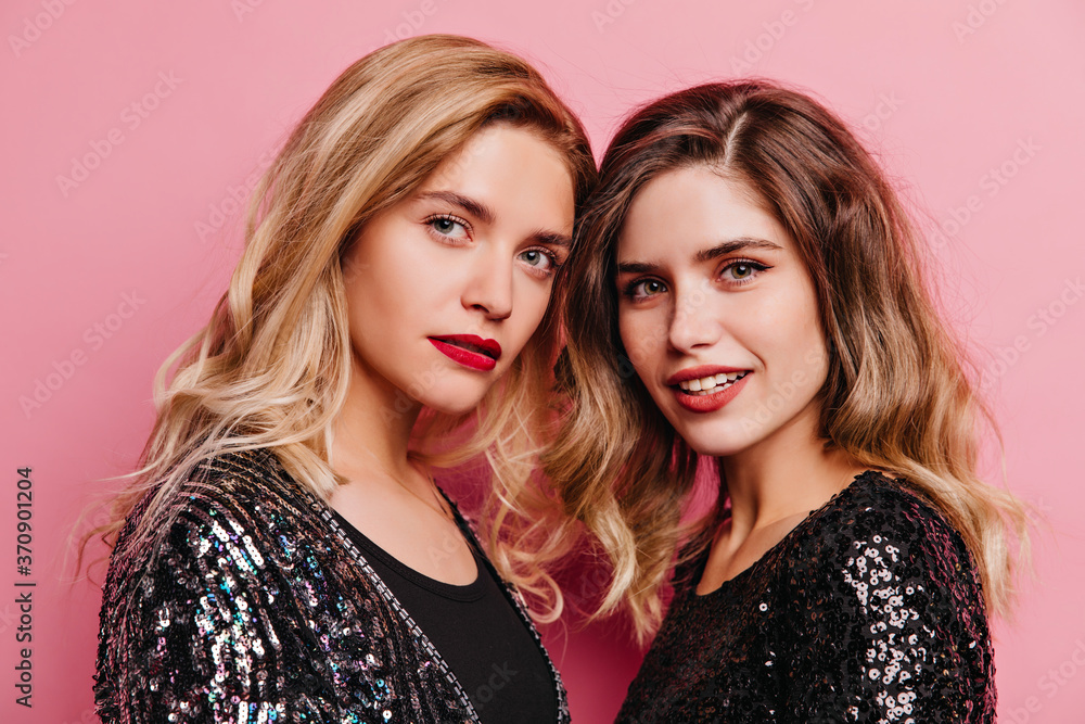 Two adorable girls posing with gently smile on pink background. Pleased female models looking to camera. Two adorable girls posing with gently smile on pink background. Pleased female models looking to camera.
