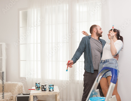 Couple dancing during home repair using brushes dipped in blue paint. Hipster renovating his house. Apartment redecoration and home construction while renovating and improving. Repair and decorating.