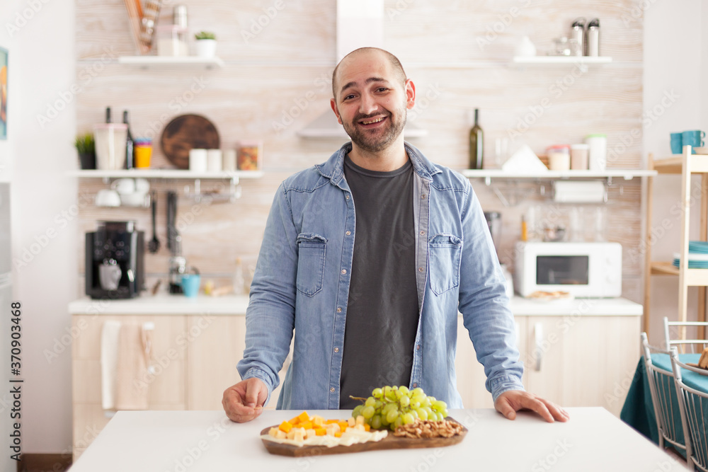 Cheerful young man in kitchen smiling looking at at camera with various cheeseboard and grapes on the table. Guy with assorted and nutritious appetizier.