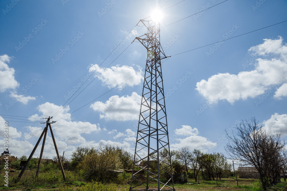 A metal high-voltage tower against a blue sky with white clouds towering above the trees. Power supply