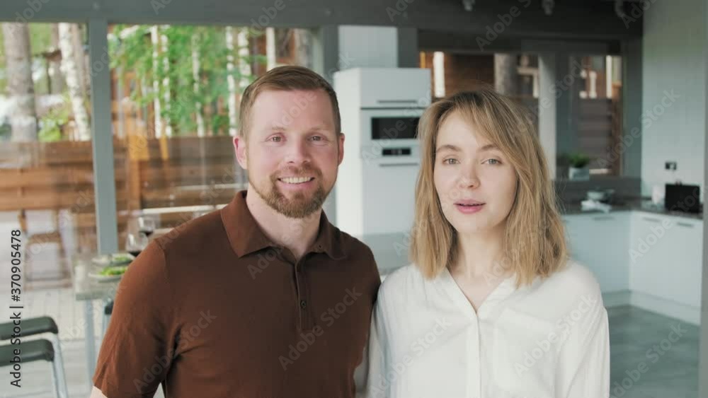 Tracking portrait of attractive young couple smiling for camera in kitchen of modern house with panoramic windows Tracking portrait of attractive young couple smiling for camera in kitchen of modern house with panoramic windows