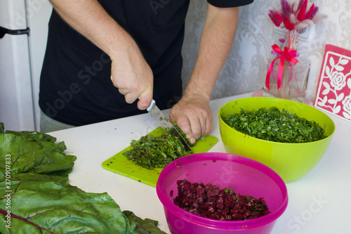 The man cut the tops of beets on a cutting Board. Next to it on the surface of the table are parsley and beet tops. In the container, crushed parsley and beet tops.