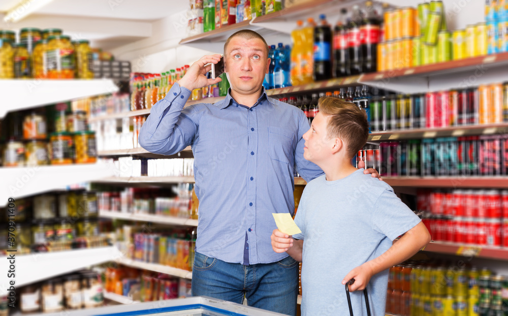 positive young man talking on phone while visiting supermarket with preteen son
