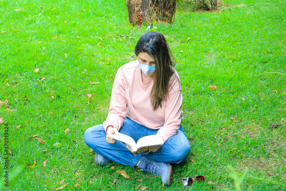Caucasian woman with a mask due to covid-19 reading a book on the grass in the park Caucasian woman with a mask due to covid-19 reading a book on the grass in the park