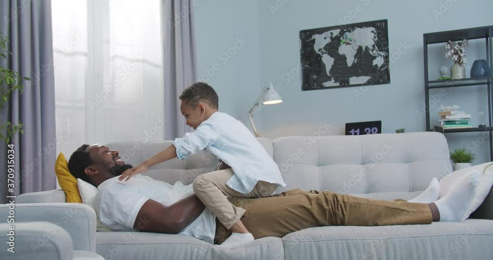 Handsome joyful African American man having fun and lifting small son up like flying in living room at home. Cheerful father playing with cute happy little boy lying on sofa indoors. Family concept Handsome joyful African American man having fun and lifting small son up like flying in living room at home. Cheerful father playing with cute happy little boy lying on sofa indoors. Family concept