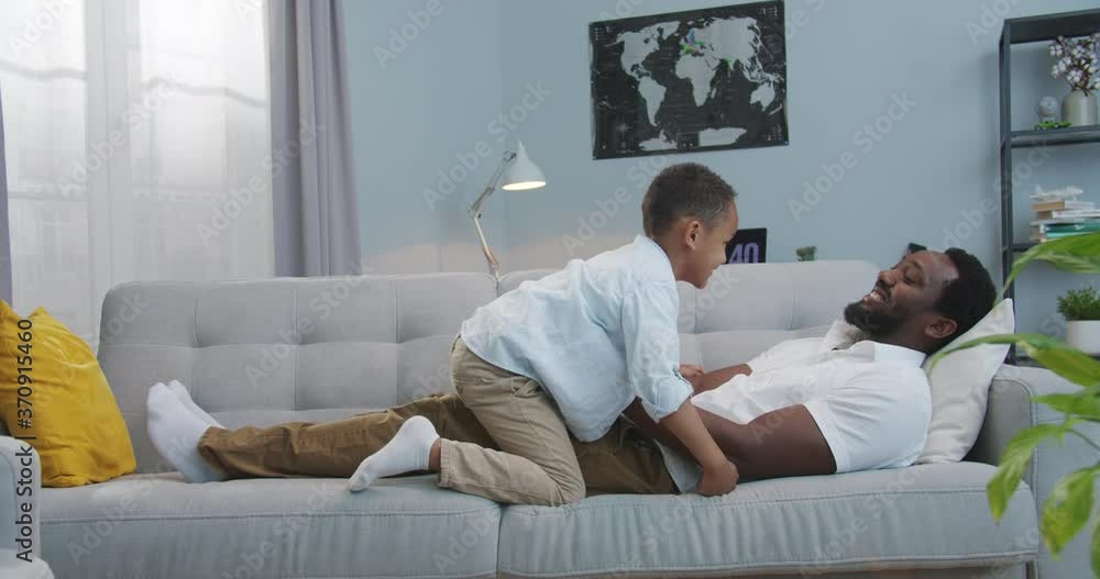 Handsome happy African American father having fun and lifting small son up like flying plane in living room. Joyful handsome man playing with cute little boy lying on sofa at home. Leisure concept Handsome happy African American father having fun and lifting small son up like flying plane in living room. Joyful handsome man playing with cute little boy lying on sofa at home. Leisure concept