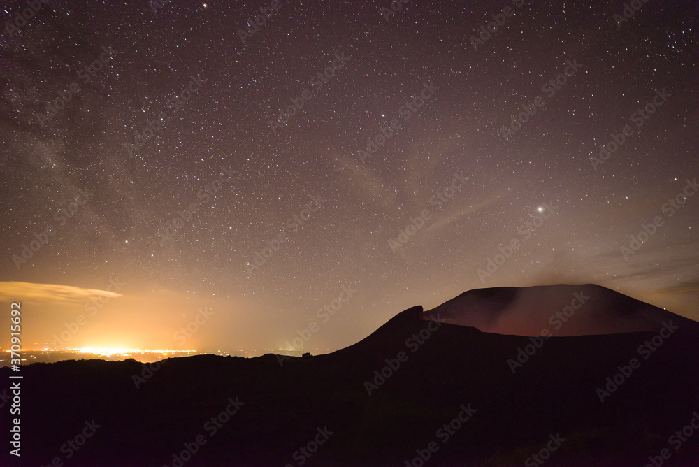 scenic panorama of the active volcano Telica with sulfuric smoke coming out of the volcanic cone at night with starry sky and lights of the city of León in the background