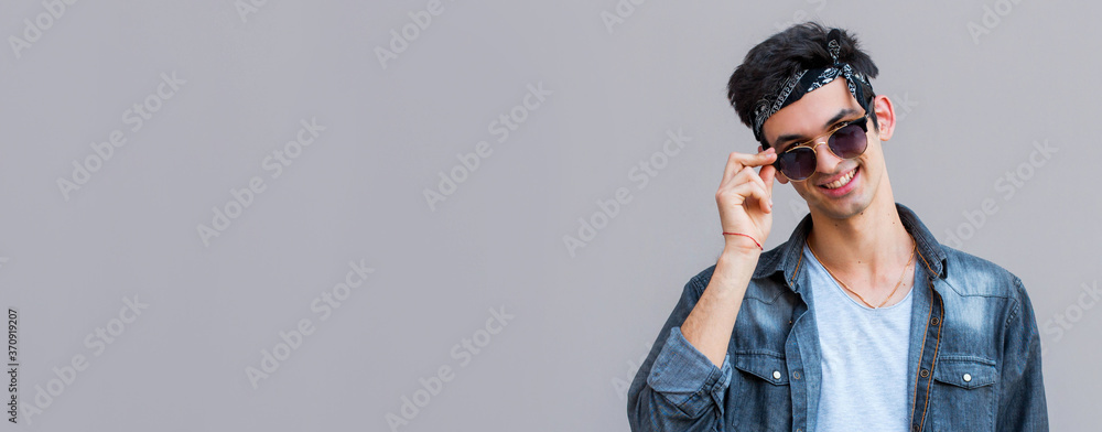 Confident in his perfect style. Handsome young fashionable man adjusting his sunglasses and looking at camera while standing against gray background Confident in his perfect style. Handsome young fashionable man adjusting his sunglasses and looking at camera while standing against gray background