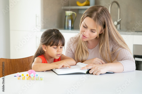 Pretty mom reading book with daughter in kitchen. Cute girl in red shirt pointing with finger in text, sitting and learning with young blonde mother. Family time, childhood and education concept