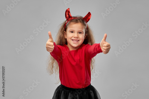 halloween  holiday and childhood concept - smiling girl in party costume and red devil s horns over grey background