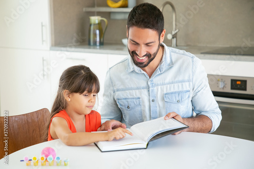 Bearded dad reading book with daughter in kitchen. Cute girl in red shirt pointing with finger in text, sitting and learning with young handsome father. Family time, childhood and education concept
