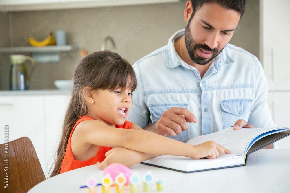 Lovely cute girl pointing at text and talking with bearded dad. Handsome young father teaching little daughter and reading book with her at kitchen table. Family time, childhood and education concept Lovely cute girl pointing at text and talking with bearded dad. Handsome young father teaching little daughter and reading book with her at kitchen table. Family time, childhood and education concept