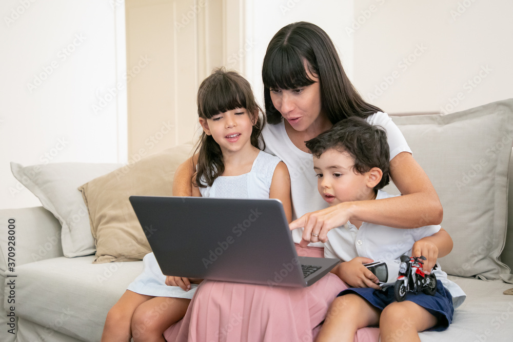 Cheerful excited mom hugging happy kids and pointing at laptop screen. Family sitting on couch at home and watching movie. Medium shot. Communication or entertainment concept Cheerful excited mom hugging happy kids and pointing at laptop screen. Family sitting on couch at home and watching movie. Medium shot. Communication or entertainment concept