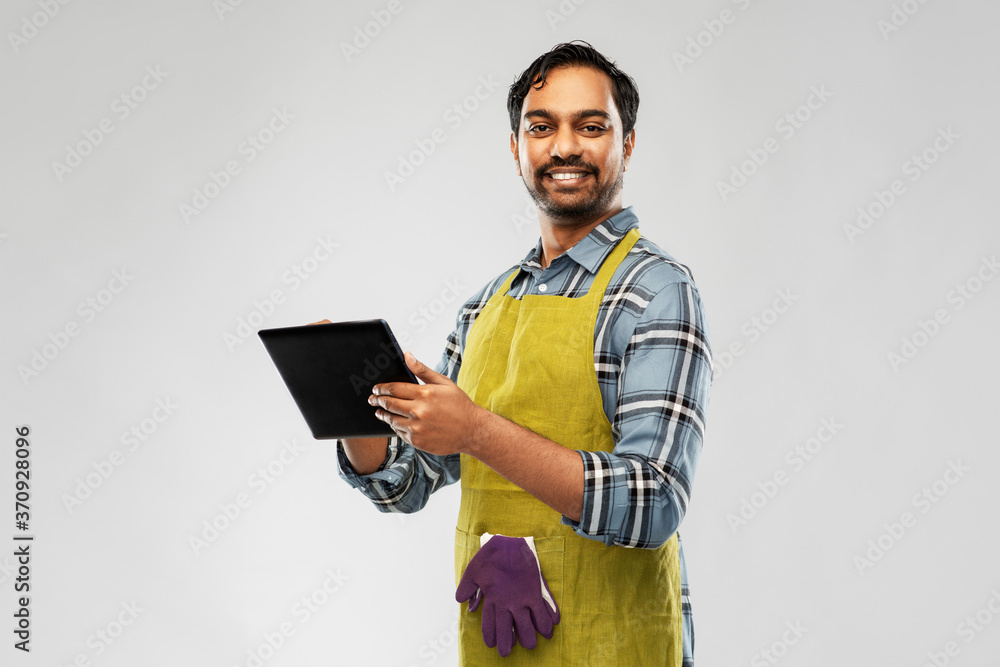 gardening, farming and technology concept - happy smiling indian male gardener or farmer in apron with tablet pc computer over grey background gardening, farming and technology concept - happy smiling indian male gardener or farmer in apron with tablet pc computer over grey background