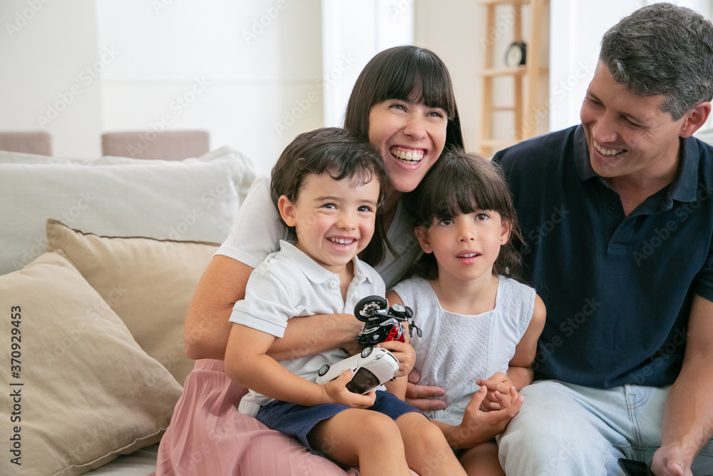 Cheerful funny parents and two kids watching funny movie at home, sitting on couch in living room and looking away and laughing. Medium shot. Leisure time with family or television concept Cheerful funny parents and two kids watching funny movie at home, sitting on couch in living room and looking away and laughing. Medium shot. Leisure time with family or television concept