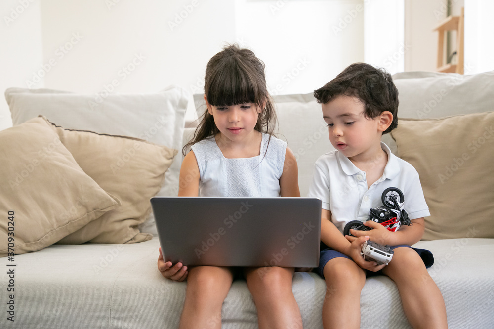 Focused pensive little brother and sister sitting on sofa at home, using laptop for video call or watching video or movie. Front view. Kids leisure time or internet concept Focused pensive little brother and sister sitting on sofa at home, using laptop for video call or watching video or movie. Front view. Kids leisure time or internet concept