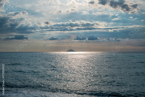promienie słoneczne przebijające się przez chmury. W oddali widać aktywny wulkan Stromboli