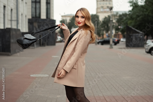 Portrait of young beautiful woman with long blonde hair in beige trench coat with a bag on the city street