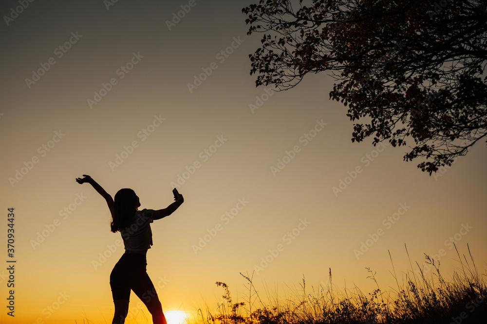 The silhouette of a single woman with hand up using a selfie to take a picture of herself at sunset. Beautiful sky at sunset. Copy space.