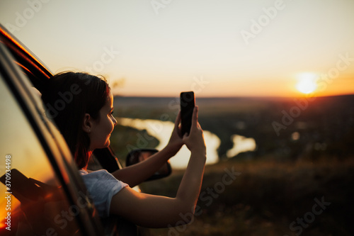 Obraz na płótnie Young woman traveler inside car, looking and taking a photo beautiful sunset. w salonie