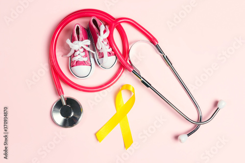 Childhood Cancer Awareness Golden Ribbon with blue baby sneakers and stethoscope on pink background with copy space.