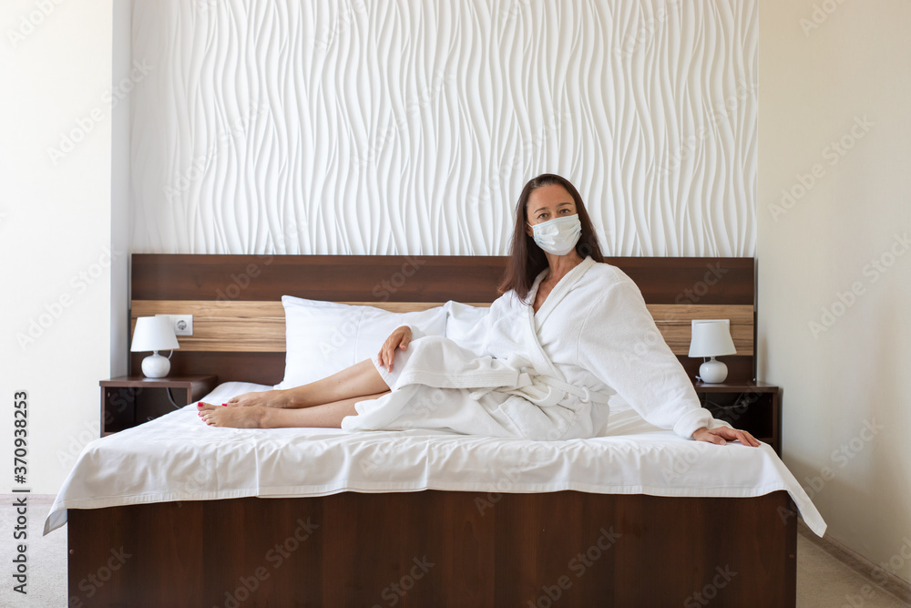 Vacation during quarantine and coronavirus, a middle-aged woman in a medical face mask and white bathrobe sitting on bed in hotel room. Copy space Vacation during quarantine and coronavirus, a middle-aged woman in a medical face mask and white bathrobe sitting on bed in hotel room. Copy space
