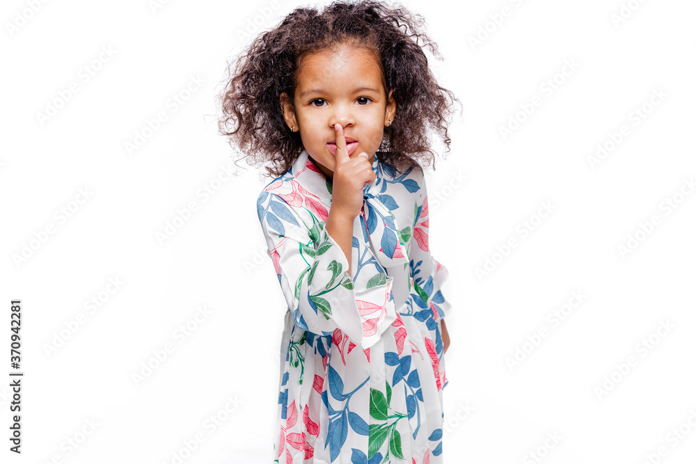 Cheerful little african girl wearing white floral dress standing isolated over white background, showng silence gesture Cheerful little african girl wearing white floral dress standing isolated over white background, showng silence gesture