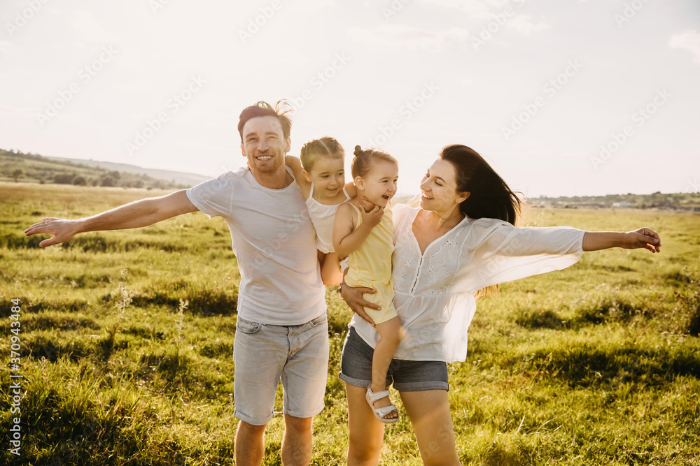 Fototapeta premium Family of four playing in a field on a summer day. Mother, father and two daughters running on green grass, pretending to be airplanes.