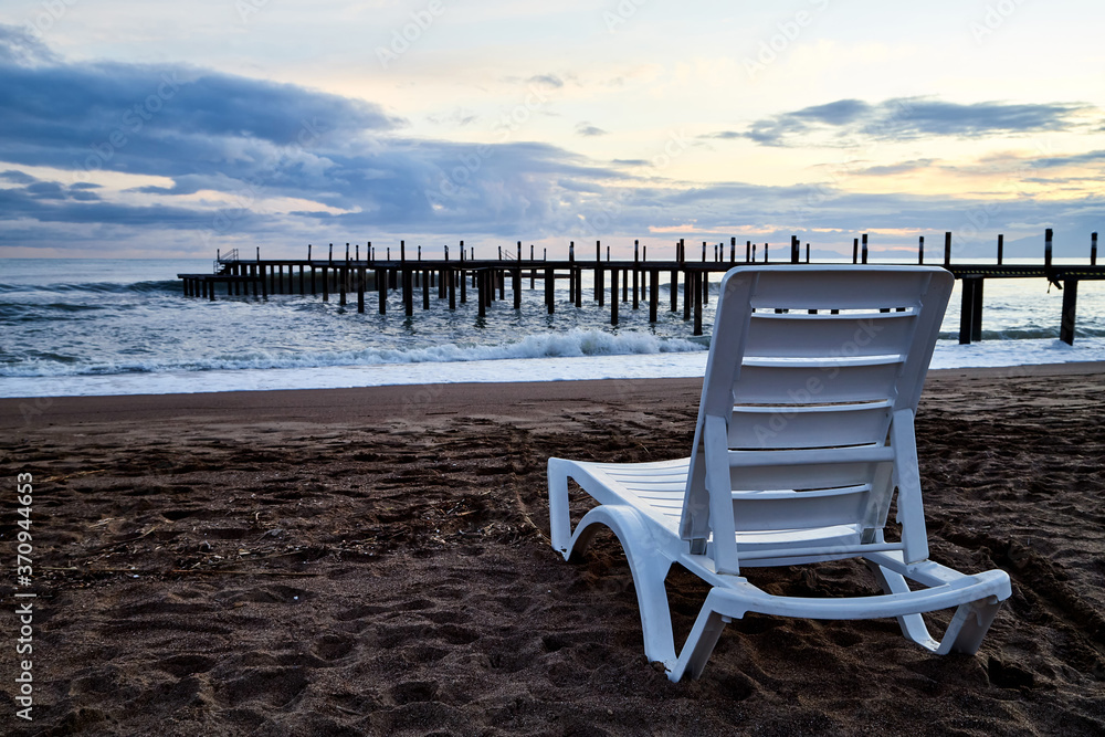 Sunbed on the sand beach and view to water of sea, waves with white foam, pierce and sky with clouds in a nice evening.