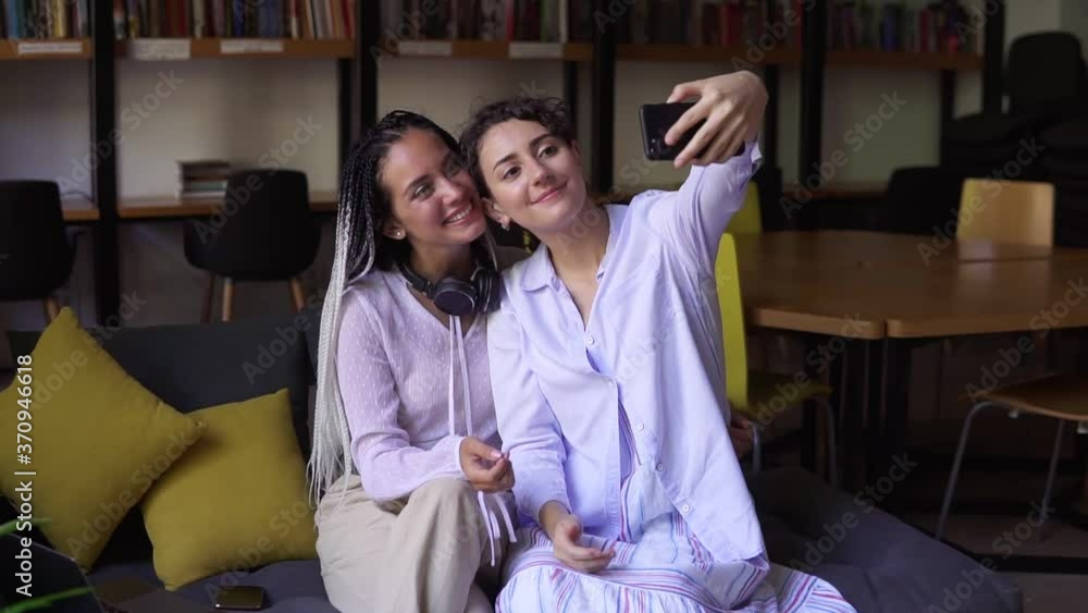 Young female students sitting on sofa in library surrounded by books. Girl is holding her smartpone and taking selfie with her university friend with stylish dreadlocks and headphones on neck Young female students sitting on sofa in library surrounded by books. Girl is holding her smartpone and taking selfie with her university friend with stylish dreadlocks and headphones on neck