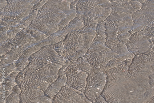  Auf das flache Wasser am Ufer des Meeres kommen kleine Wellen. Von der Sonne bestrahlt, produzieren sie viele kleine Schatten auf dem Sand des Meeres Bodens.