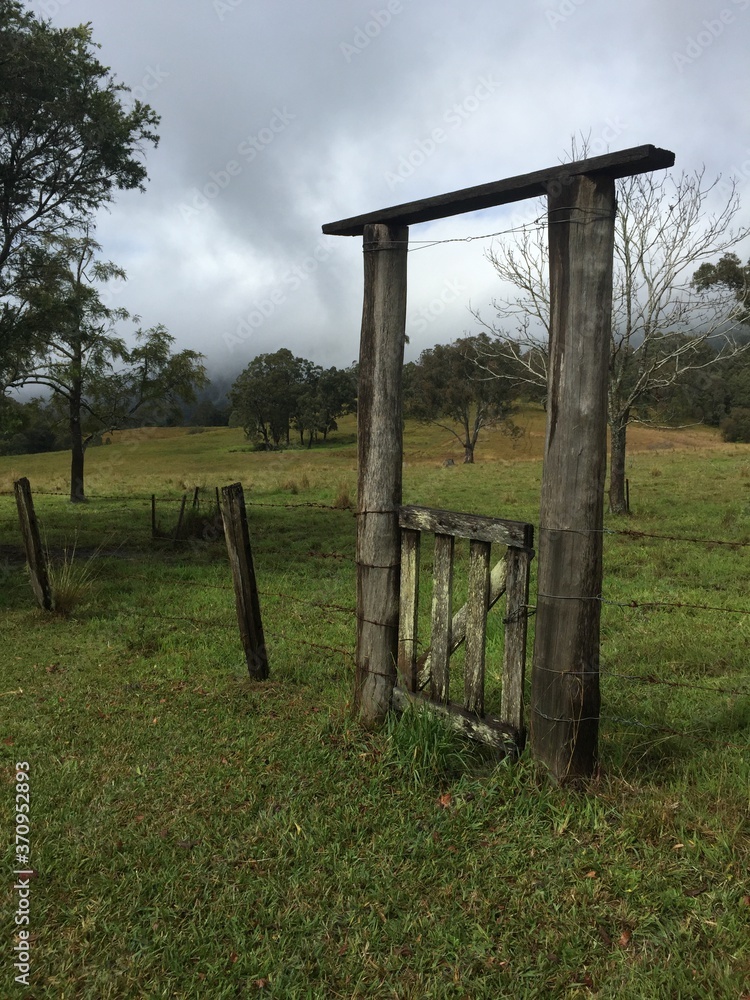 Old wooden gate through a barbed wire fence in the the countryside with green grass, trees and an overcast sky. 