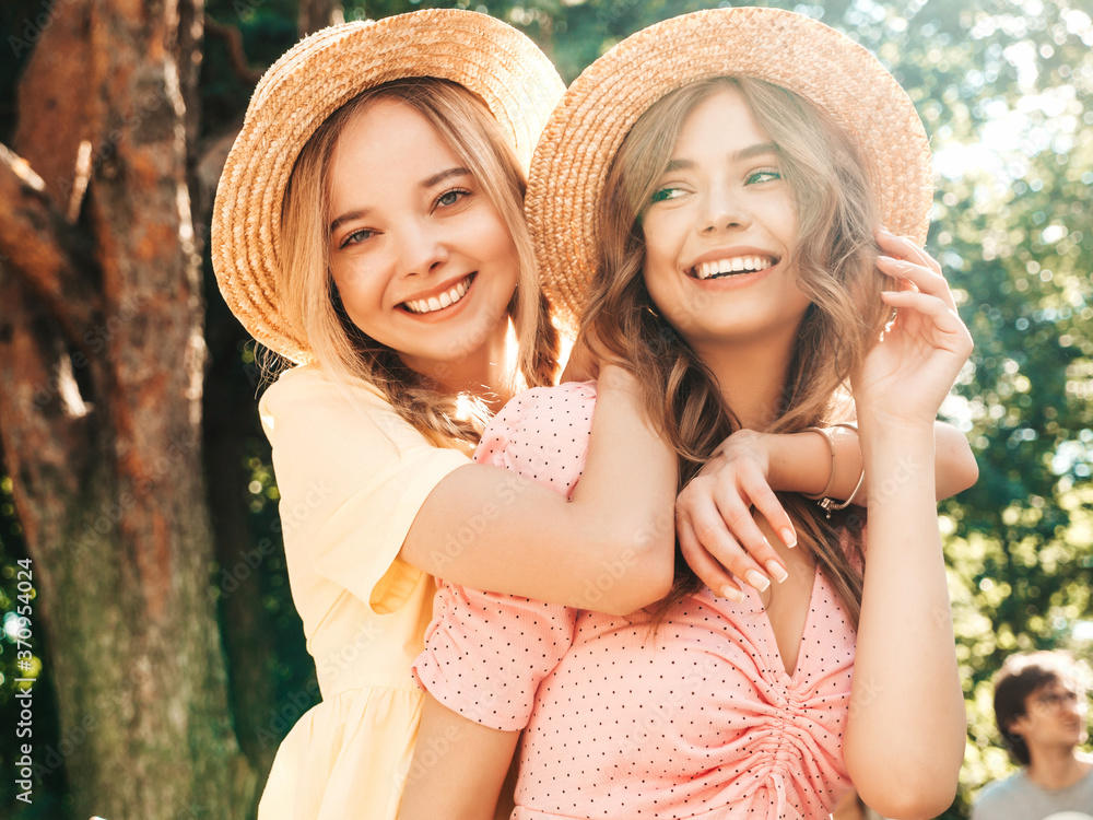 Two young beautiful smiling hipster woman in trendy summer sundress. Sexy carefree women posing in the park in hats. Positive models having fun and hugging at sunset