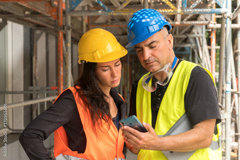 Two workers on construction site wearing safety vest, helmet and goggles checking the work plan on a digital tablet computer Two workers on construction site wearing safety vest, helmet and goggles checking the work plan on a digital tablet computer