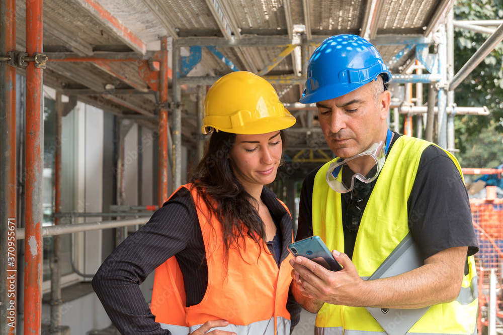 Two workers on construction site wearing safety vest, helmet and goggles checking the work plan on a digital tablet computer Two workers on construction site wearing safety vest, helmet and goggles checking the work plan on a digital tablet computer