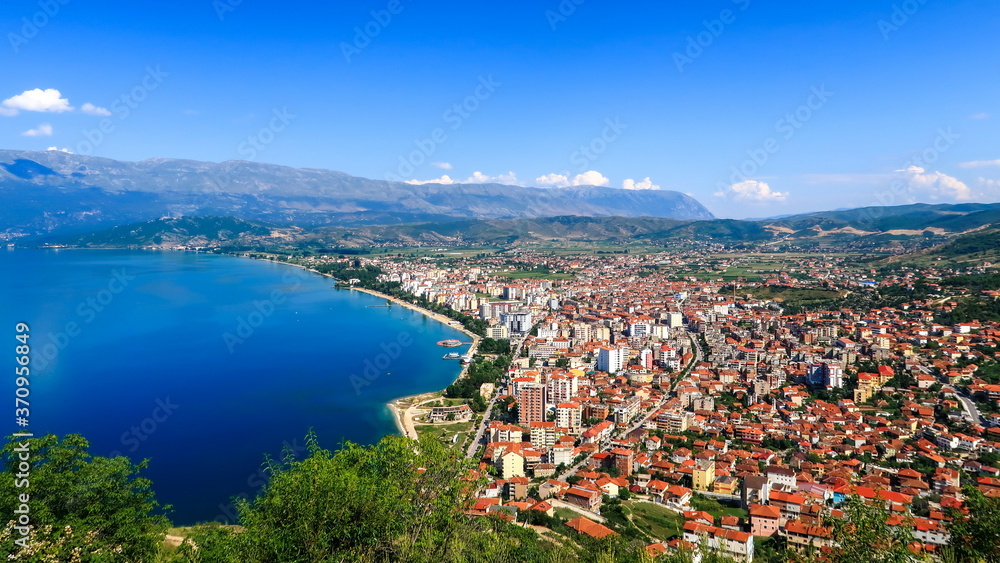 City buildings with red roofs at the shore of a lake with mountains in the background. View of Pogradec city and Ohrid lake, Pogradec, Albania