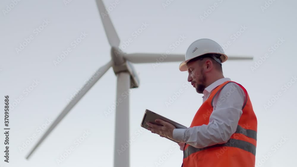 An electrical engineer looks at a modern digital tablet and raises his hand with an orange battery against the background of a windmill. An engineer with a beard, wearing a helmet and vest, holds a An electrical engineer looks at a modern digital tablet and raises his hand with an orange battery against the background of a windmill. An engineer with a beard, wearing a helmet and vest, holds a