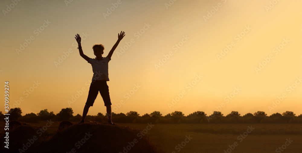 Silhouette of a happy jumping child-boy, over a haystack on the background of a field with a beautiful sunset.The concept of a carefree childhood. Beautiful summer landscape, copyspace, banner