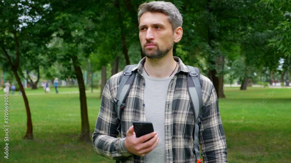 Gray-haired, focused man travel blogger records video sightseeing on smartphone for social media subscribers. Guy tourist in shirt, backpack walks in park makes photos on smart phone. Slow motion Gray-haired, focused man travel blogger records video sightseeing on smartphone for social media subscribers. Guy tourist in shirt, backpack walks in park makes photos on smart phone. Slow motion