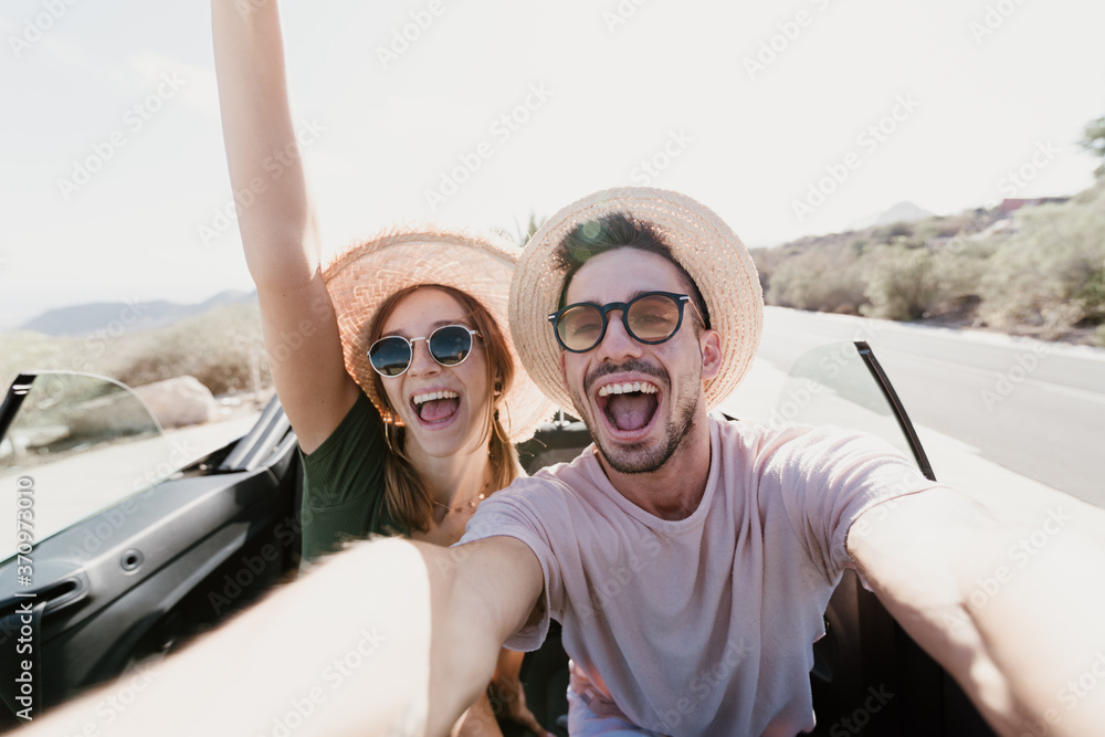 Happy excited couple taking a selfie driving on a convertible car at holidays. Young people at vacation on a rent car.