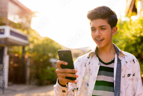 Happy young handsome Indian teenage boy using phone outdoors