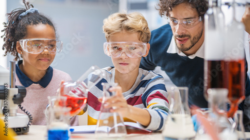 Elementary School Science Classroom: Enthusiastic Teacher Helps with Chemistry to Diverse Group of Children, Little Boy Mixes Chemicals in Beakers. Children Learn with Interest