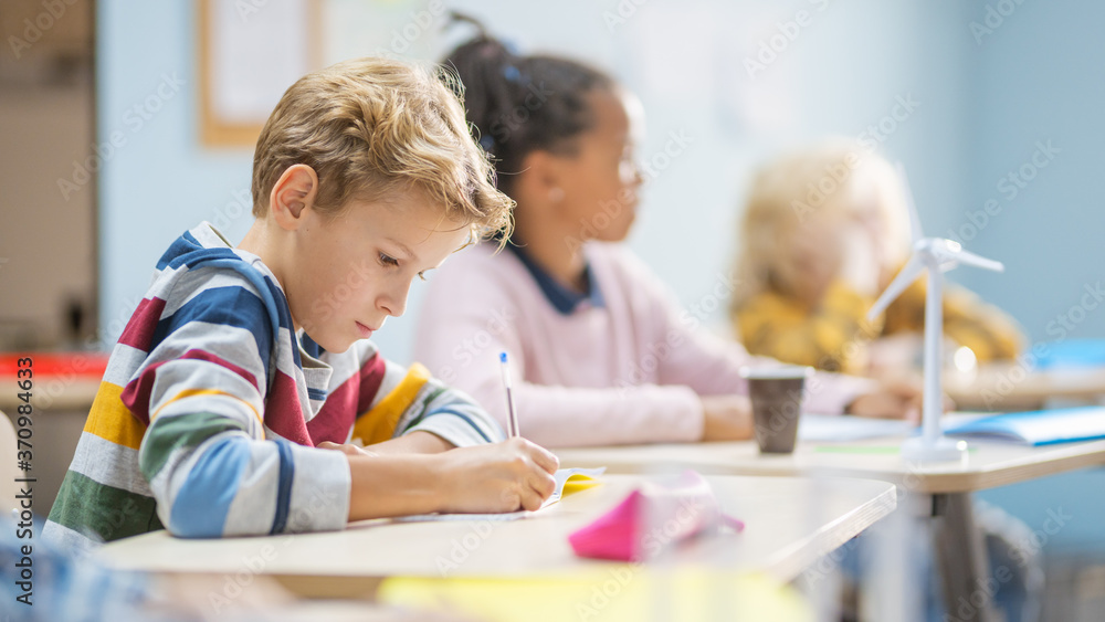 In Elementary School Classroom Brilliant Caucasian Boy Writes in Exercise Notebook, Taking Test and Writing Exam. Junior Classroom with Group of Children Working Diligently and Learning New Stuff In Elementary School Classroom Brilliant Caucasian Boy Writes in Exercise Notebook, Taking Test and Writing Exam. Junior Classroom with Group of Children Working Diligently and Learning New Stuff