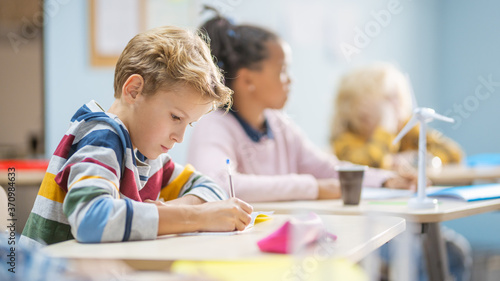In Elementary School Classroom Brilliant Caucasian Boy Writes in Exercise Notebook, Taking Test and Writing Exam. Junior Classroom with Group of Children Working Diligently and Learning New Stuff