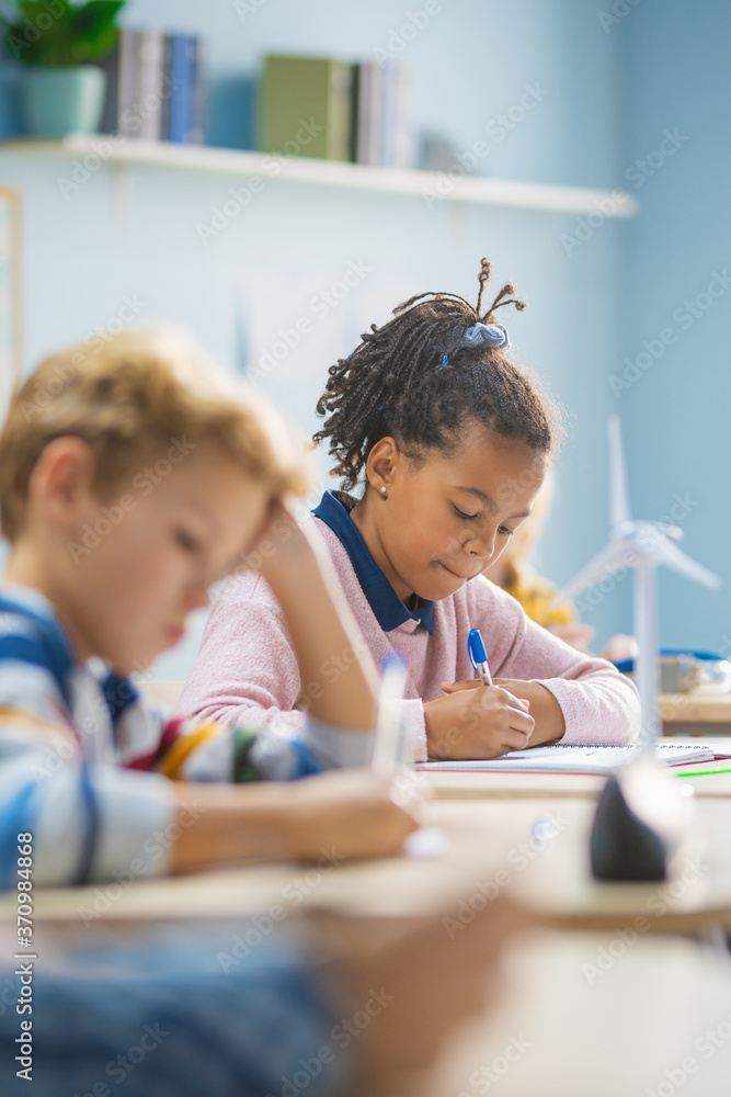 In Elementary School Classroom Brilliant Black Girl Writes in Exercise Notebook, Taking Test and Writing Exam. Junior Classroom with Group of Bright Children Working Diligently and Learning New Stuff In Elementary School Classroom Brilliant Black Girl Writes in Exercise Notebook, Taking Test and Writing Exam. Junior Classroom with Group of Bright Children Working Diligently and Learning New Stuff