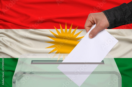 male voter lowers the ballot in a transparent ballot box against the background of the national flag of Kurdistan, rojava, concept of state elections, referendum